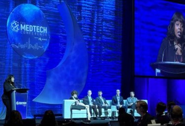 Panel discussion at The MedTech Conference, featuring speakers seated on stage with a large blue backdrop displaying the event logo. A moderator stands at a podium on the left, with a screen displaying a speaker in the background.