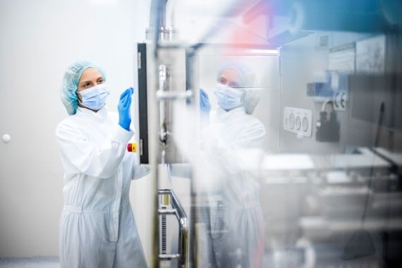 A woman wearing proper safety clothes seen taping on the screen of a big complex machine in pharmaceutical industry while her reflection is visible on the machine.