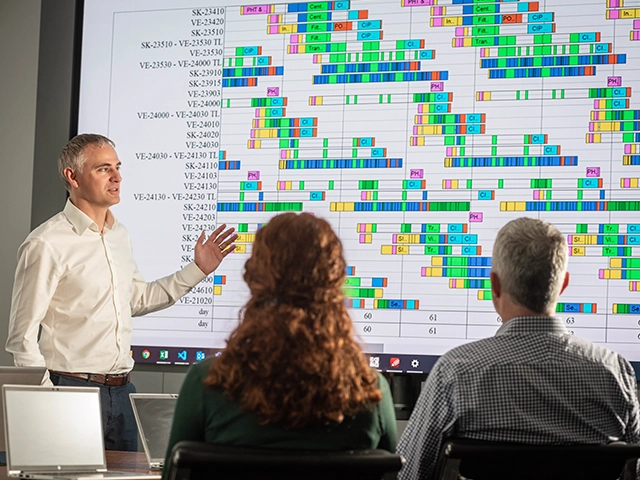 A man presenting from a large screen filled with data, presenting to a man and a woman