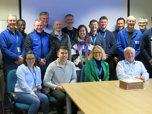 Group photo of team celebrating with a cake in a meeting room.