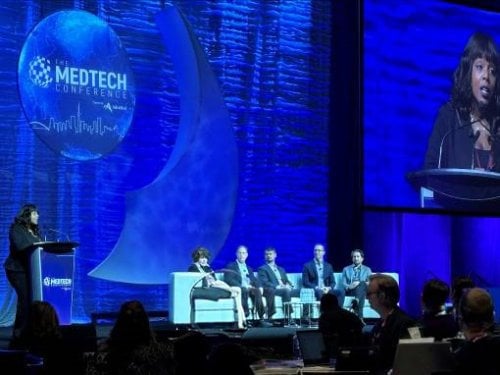 Panel discussion at The MedTech Conference, featuring speakers seated on stage with a large blue backdrop displaying the event logo. A moderator stands at a podium on the left, with a screen displaying a speaker in the background.