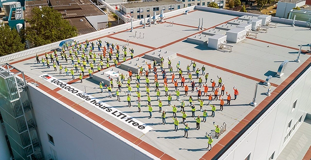 Group of people waving at a camera while standing on a building roof, wearing full safety gear